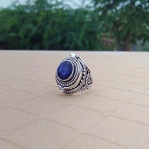 Peut inclure: Une bague en argent avec une pierre précieuse bleue sertie dans une bélière ovale décorative. La bague présente des détails complexes et un design à charnière.