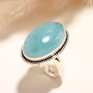 Puede incluir: Un anillo de plata de ley con una gran piedra preciosa cabujón de forma ovalada, lisa y de color azul claro. El engaste está enmarcado por un borde de metal trenzado tipo cuerda. La banda interior del anillo tiene el sello 925 de plata.