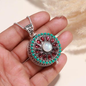 Op de afbeelding: Een zilveren hangerketting met een rond, sierlijk ontwerp. De hanger heeft een centrale witte steen, omringd door rode en turkooizen stenen. De ketting wordt in een hand gehouden.