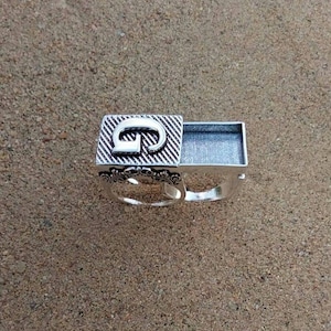Può includere: Un anello d'argento con una scatola incernierata sulla parte superiore. La scatola è aperta e la parte superiore ha la lettera "G" incisa.