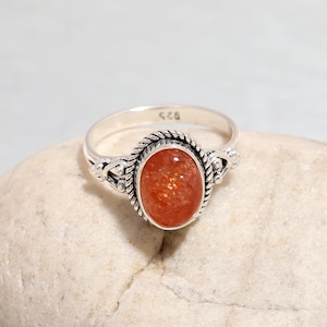 Puede incluir: Un anillo de plata con una piedra preciosa de piedra de sol roja engastada en un diseño de cuerda trenzada. El anillo está marcado con "925" en la banda interior.