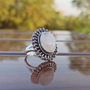 Könnte beinhalten: Ein silberner Ring mit einem großen ovalen weißen Edelstein, der in einer detaillierten silbernen Fassung sitzt. Der Ring hat ein dekoratives Design mit einem kreisförmigen Muster um den Stein.