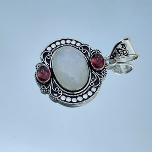 Puede incluir: Un colgante de plata con una gran piedra de luna ovalada y dos piedras rojas m&aacute;s peque&ntilde;as. El colgante tiene un dise&ntilde;o intrincado con espirales y puntos.