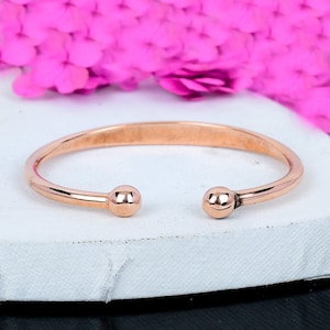 May include: A rose gold open cuff bracelet with a minimalist design. The bracelet features a smooth, rounded band that curves to end with a small, spherical ball on each end. The bracelet is displayed on a white surface with pink flowers in the background.