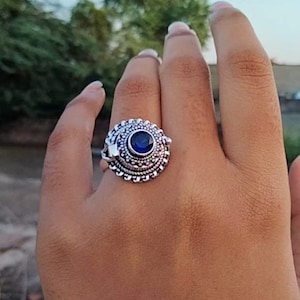 May include: A silver-colored ring with an ornate design. The ring features a large, round, dark blue stone in the center, surrounded by a detailed, textured pattern. The ring is on a hand, with the background blurred.
