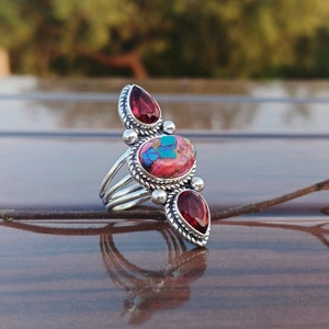 Könnte beinhalten: Ein silberner Ring mit drei Steinen. Der mittlere Stein ist ein ovaler Stein in Pink und Blau, und die beiden seitlichen Steine sind rote tropfenförmige Steine.