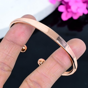 Puede incluir: Una pulsera de brazalete de oro rosa con un acabado liso y pulido. La pulsera tiene un diseño simple y elegante y está sujeta por una mano. El fondo es blanco y negro con flores rosas.