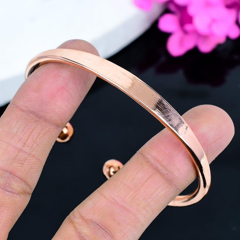 May include: A rose gold cuff bracelet with a smooth, polished finish. The bracelet has a simple, elegant design and is held by a hand. The background is black and white with pink flowers.