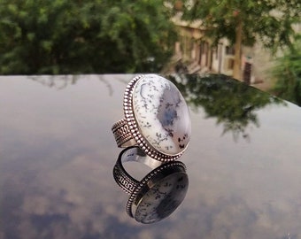 Può includere: Un anello d'argento con una gemma ovale bianca con motivi grigi e neri. L'anello ha una fascia decorativa e un bordo di perline attorno alla pietra. L'anello è esposto su una superficie riflettente.