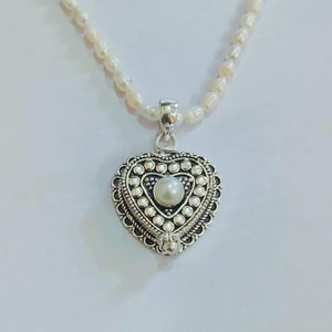 Puede incluir: Un colgante de plata en forma de corazón con una perla en el centro, colgando de un collar de perlas blancas.