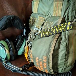 May include: An olive green flight bag with orange accents and the text "PULL TO EJECT" on a yellow and black tag. A pair of green aviation headsets and a black glove are also visible. The bag has a military aesthetic.