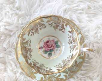 May include: A vintage teacup and saucer set with a delicate floral design. The cup and saucer are both light green with gold accents and a pink rose in the center.