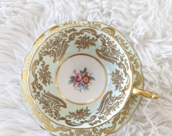 Puede incluir: Un juego de taza y platillo de té verde y dorado con un diseño floral. El platillo tiene un borde festoneado. La taza de té tiene un asa dorada. El juego está marcado con "Paragon Double Warrant 1939-49".