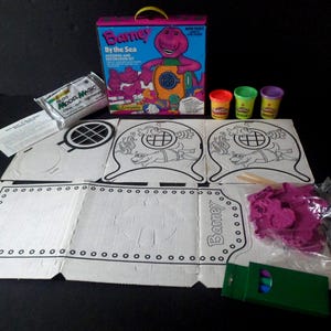 May include: A Barney By the Sea molding and decorating kit including a cardboard box, three small containers of Play-Doh, a bag of Model Magic, a green crayon box, a plastic bag of purple plastic shapes, and several white cardboard cutouts with black outlines.
