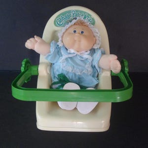 May include: A Cabbage Patch Kids doll with blonde hair and blue eyes, wearing a light blue dress with white lace trim and a matching bonnet. The doll is seated in a cream-colored plastic carrier with a green handle and a green logo at the top.