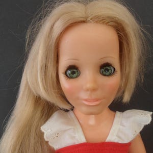 May include: A vintage doll with long, blonde hair and large, green eyes. The doll has a fair complexion, rosy cheeks, and a small, smiling mouth. She wears a red top with a white, ruffled collar.
