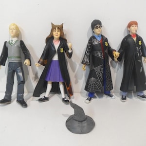 May include: Four plastic figurines of characters from a fantasy series, including a blonde-haired figure in a gray vest and black pants, a figure in a purple skirt and black robe, and two figures in black robes. A gray hat is in front.