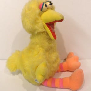 May include: A plush toy of Big Bird from Sesame Street. The yellow bird has a large beak, orange feet, and striped legs. The toy is sitting down with its mouth open, revealing a red interior.