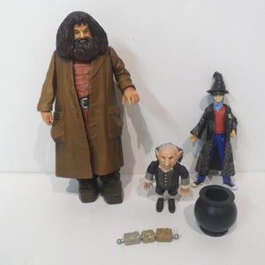 May include: A collection of Harry Potter character figurines, including Hagrid in a brown coat, Harry Potter in a black robe, and a goblin figure. Also included are a black cauldron and a small, beaded accessory.