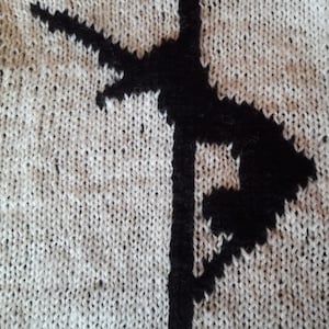 May include: Close-up of a knitted fabric with a black design. The design is a stylised letter F, with a small circle at the top.