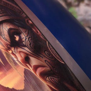 Puede incluir: Un primer plano de una ilustración fantástica de una persona con intrincados tatuajes faciales y un tocado. La cara de la persona está pintada con una mezcla de colores rojo, marrón y negro. El fondo es una imagen borrosa de una puesta de sol.