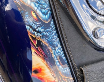 May include: Close-up of a motorcycle with a detailed dragon graphic. The design features a blue and orange dragon eye and scales, with a dark blue background. The motorcycle has a black leather seat and chrome accents.