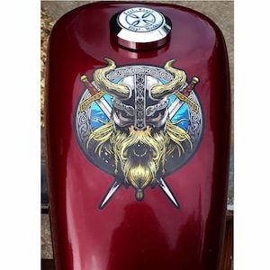Op de afbeelding: Een rode motorfiets benzinetank met een ronde sticker met daarop een Viking krijger met een gehelmde helm, een baard en gekruiste zwaarden. De sticker heeft een blauw en gouden kleurenschema met een Keltische knoop rand.
