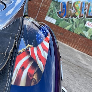 May include: A close-up of a motorcycle with a blue and purple paint job. The motorcycle has a graphic of an eagle holding an American flag on the side. The motorcycle is parked in front of a brick building with a mural that says "Jasper, Alabama".