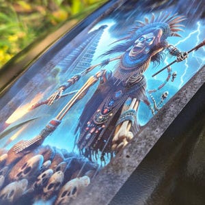 Puede incluir: Un gráfico azul y negro de una figura esquelética que lleva un tocado y sostiene una antorcha en llamas. La figura está de pie frente a una estructura de piedra con cráneos en la base.