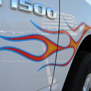 Op de afbeelding: Witte vrachtwagen met chromen "F-150" embleem en vlamstickers. De vlammen zijn blauw omlijnd, met een rood interieur en gele accenten. De band is zwart met een zilveren velg.