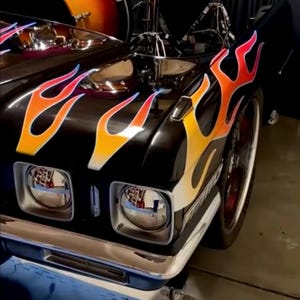 Puede incluir: Un coche negro con un diseño de llamas en naranja, amarillo y rojo. El coche tiene detalles cromados y una batería en el fondo. La batería tiene un degradado de naranja y marrón.