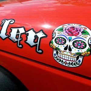 May include: A red car with a white decal that says "Day of the Dead" and a colorful sugar skull sticker.