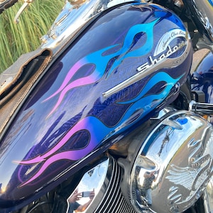May include: A blue and purple motorcycle with flames painted on the gas tank. The motorcycle has a chrome engine and a chrome eagle emblem on the side. The gas tank has a chrome emblem that says "American Classic Edition Shadow Honda".
