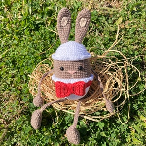 May include: A brown crocheted bunny rabbit wearing a white crocheted hat and a red bow tie. The bunny is sitting in a nest of straw.