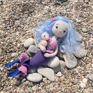 May include: A handmade crochet mermaid doll with blue hair and a purple tail, holding a smaller doll. The mermaid has pink accents and is lying on a bed of pebbles. The doll's hair is a light blue color.
