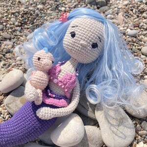May include: A handmade crochet mermaid doll with light blue hair and a pink seashell top. The mermaid is holding a smaller doll. The mermaid's tail is purple, and the doll has a light skin tone. A small pink flower is in the mermaid's hair.