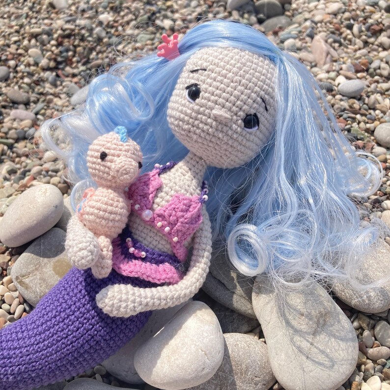 May include: A handmade crochet mermaid doll with light blue hair and a pink seashell top. The mermaid is holding a smaller doll. The mermaid's tail is purple, and the doll has a light skin tone. A small pink flower is in the mermaid's hair.