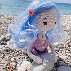 May include: A handmade crochet mermaid doll with light blue hair and a pink starfish hair clip. The doll wears a purple and pink bikini top and has a gray stone for a tail. The doll is sitting on a pebble beach.