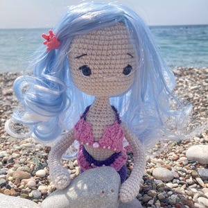 May include: A handmade crochet mermaid doll with light blue hair and a pink seashell hair clip. The doll wears a pink and purple bikini and is holding a gray stone. The doll is sitting on a pebble beach.