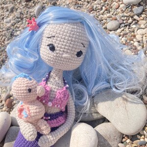 May include: A handmade crochet mermaid doll with light blue hair and a pink seashell bikini. The mermaid is holding a smaller crochet doll. The dolls are sitting on a bed of pebbles. A pink coral hair clip adorns the mermaid's hair.
