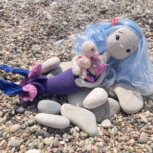 May include: A handmade crochet mermaid doll with blue hair and a purple tail, holding a smaller doll. The mermaid has a pink seashell top and a small pink crown. The dolls are resting on a bed of pebbles.