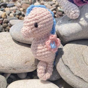 May include: A handmade crochet seahorse in soft pink with blue accents. The seahorse has a blue fin and a pink flower detail. The seahorse is sitting on a bed of gray rocks, creating a beachy aesthetic.