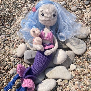 May include: A handmade crochet mermaid doll with light blue hair and a purple tail. The mermaid is holding a smaller doll. The mermaid's top is pink, and the tail has blue and pink accents. The doll is sitting on a bed of pebbles.
