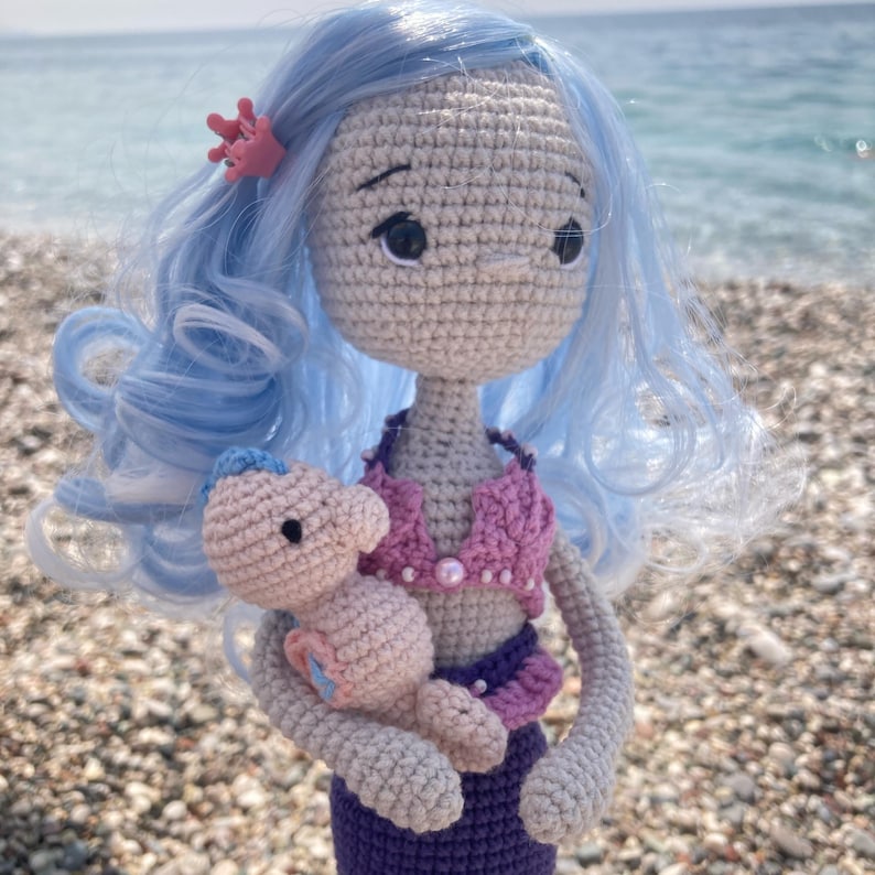 May include: A handmade crochet mermaid doll with light blue hair and a pink seashell crown. The doll wears a pink and purple top and a purple skirt. She holds a small, light pink seahorse. The doll is on a beach.