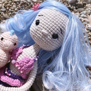 May include: Crocheted mermaid doll with light blue hair, a pink crown, and a pink and purple bikini top. A smaller crocheted doll is next to the mermaid. The dolls are handmade and have black eyes.