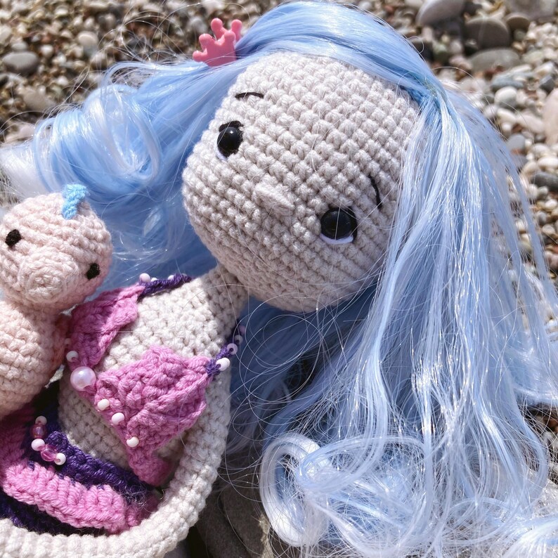 May include: Crocheted mermaid doll with light blue hair, a pink crown, and a pink and purple bikini top. A smaller crocheted doll is next to the mermaid. The dolls are handmade and have black eyes.