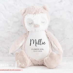 May include: A plush owl-shaped stuffed animal in soft pink and white. The owl has closed eyes and the name "Millie" is printed on the front, along with "FLOWER GIRL" and a date. A small pink heart is also present.
