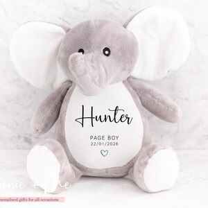May include: A gray plush elephant toy with white inner ears and feet. The elephant has the name "Hunter" printed on its belly, along with "PAGE BOY" and a date. A small green heart is also present. The toy is designed for personalized gifts.