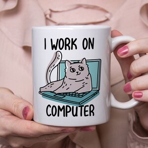 May include: White ceramic mug with a black outline of a cat sitting on a laptop. The text "I work on computer" is printed on the mug.