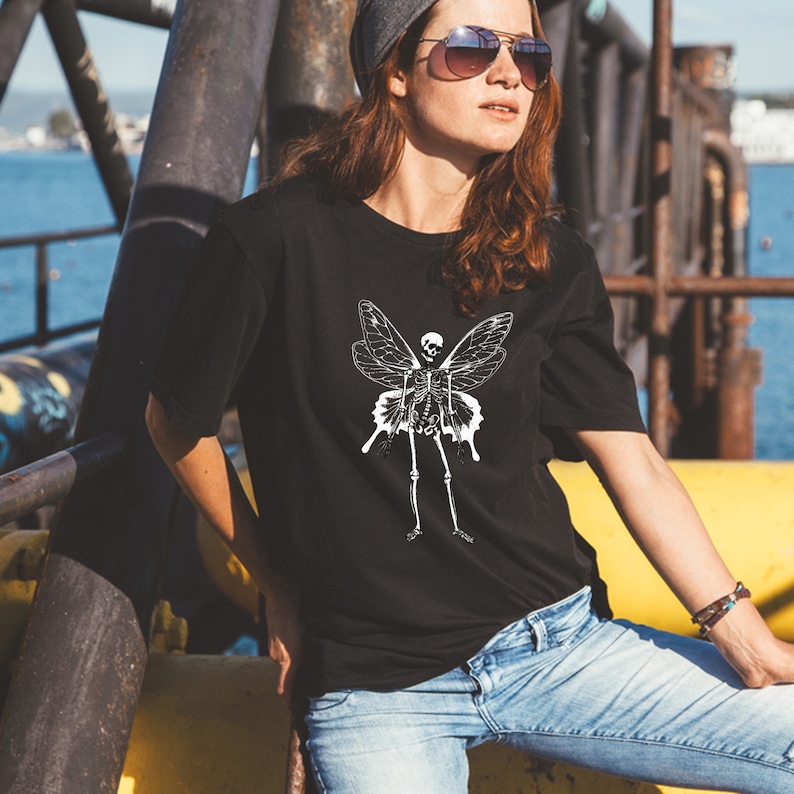 Puede incluir: Una camiseta negra con un gr&aacute;fico blanco de una hada esqueleto con alas. El hada esqueleto est&aacute; de pie con los brazos extendidos.